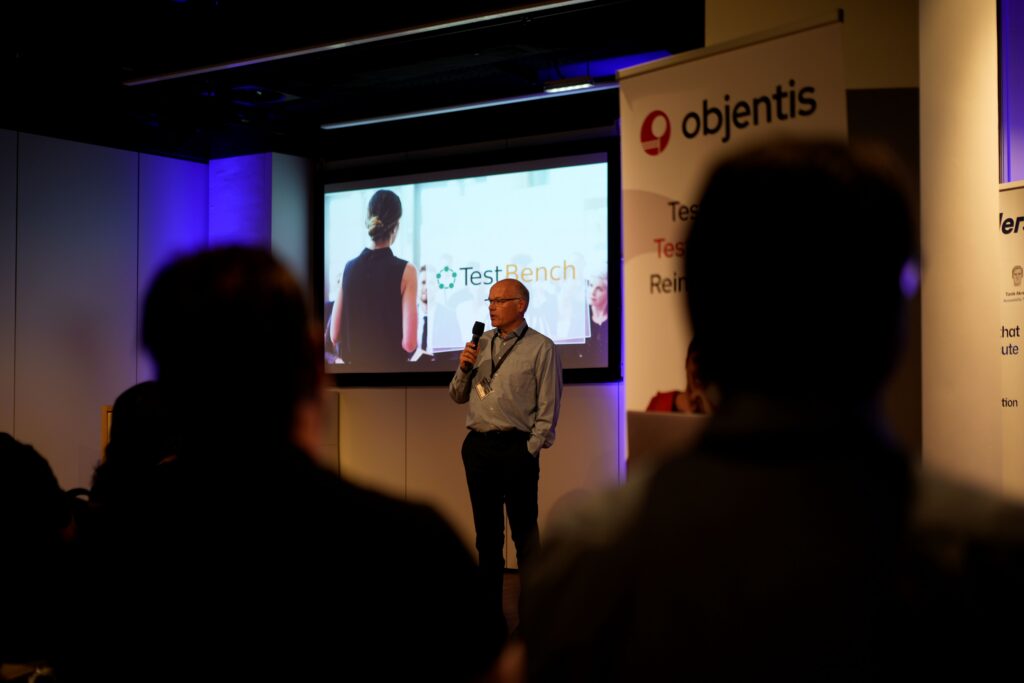 Dierk Engelhardt holding microphone stands in front of screen showing presentation about TestBench, blurred audience.